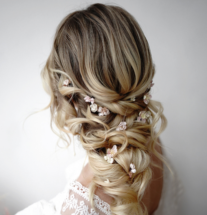 A back view of an elegant loose bridal updo with a detailed braided structure on blonde hair, featuring small and unique pink flower hair pins by @niezwykle_czesanie.