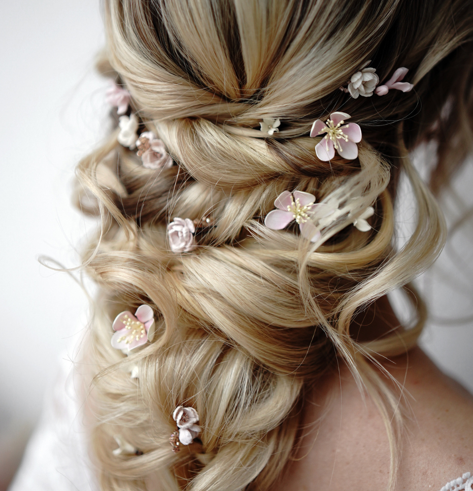 A stunning close-up of a braided updo wedding hairstyle on blonde hair, with many individual small **blush pink apple blossom flower hair pins** intricately placed throughout the delicate curls and braids.