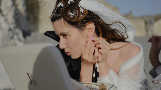 Bride getting ready by the sea, delicately putting on earrings from a keepsake DecoFlowers box, with handmade floral hair accessories in her veil