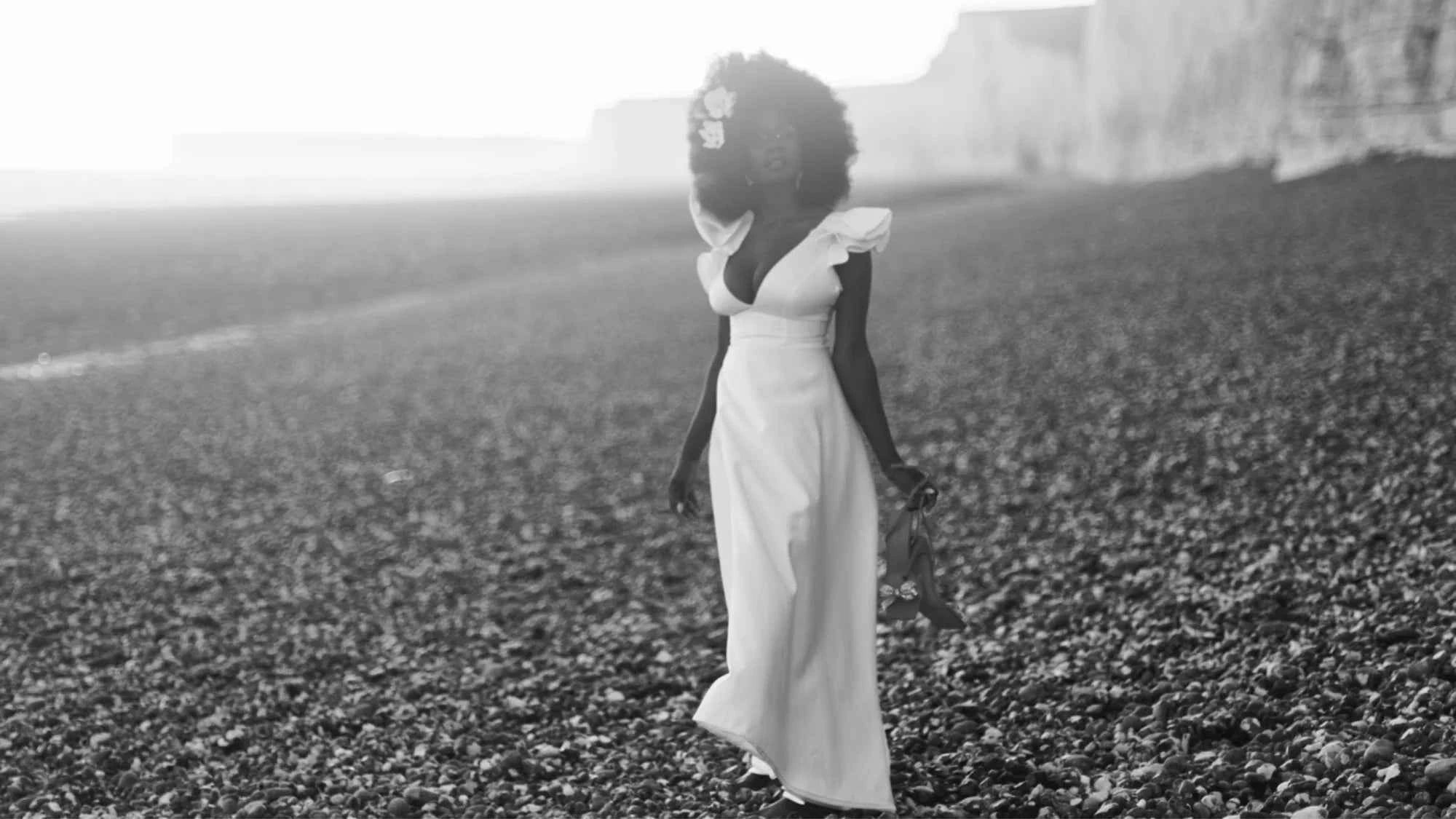Bride in a white gown walking along a pebbled beach, glowing in soft backlight with floral hair accessories, evoking serene coastal romance