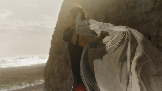 Bride in a black dress with flowing veil standing by a cliff at golden hour, capturing a sense of drama and elegance by the sea