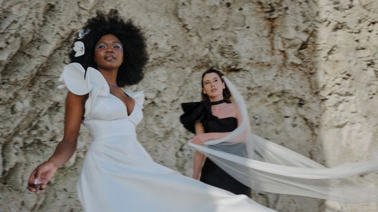 Two women standing by a cliff – one in a sculptural white bridal gown with flower makeup, the other in black with a sweeping veil – embodying contrast and modern bridal style