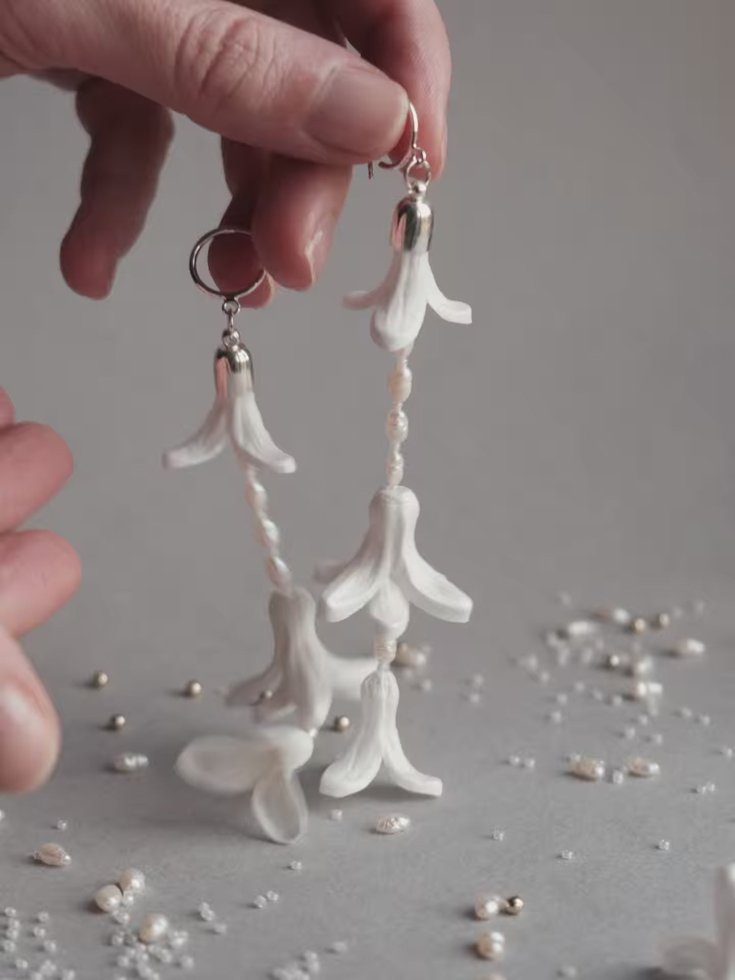 Close-up product shot of a pair of long 11cm dangling white hyacinth wedding earrings featuring intricate handmade foam petals, Japanese glass beads, and natural vintage pearls on sterling silver hooks, lying on a soft gray background.