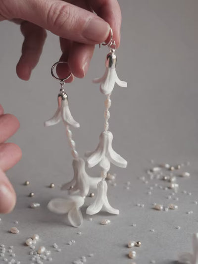 Close-up product shot of a pair of long 11cm dangling white hyacinth wedding earrings featuring intricate handmade foam petals, Japanese glass beads, and natural vintage pearls on sterling silver hooks, lying on a soft gray background.