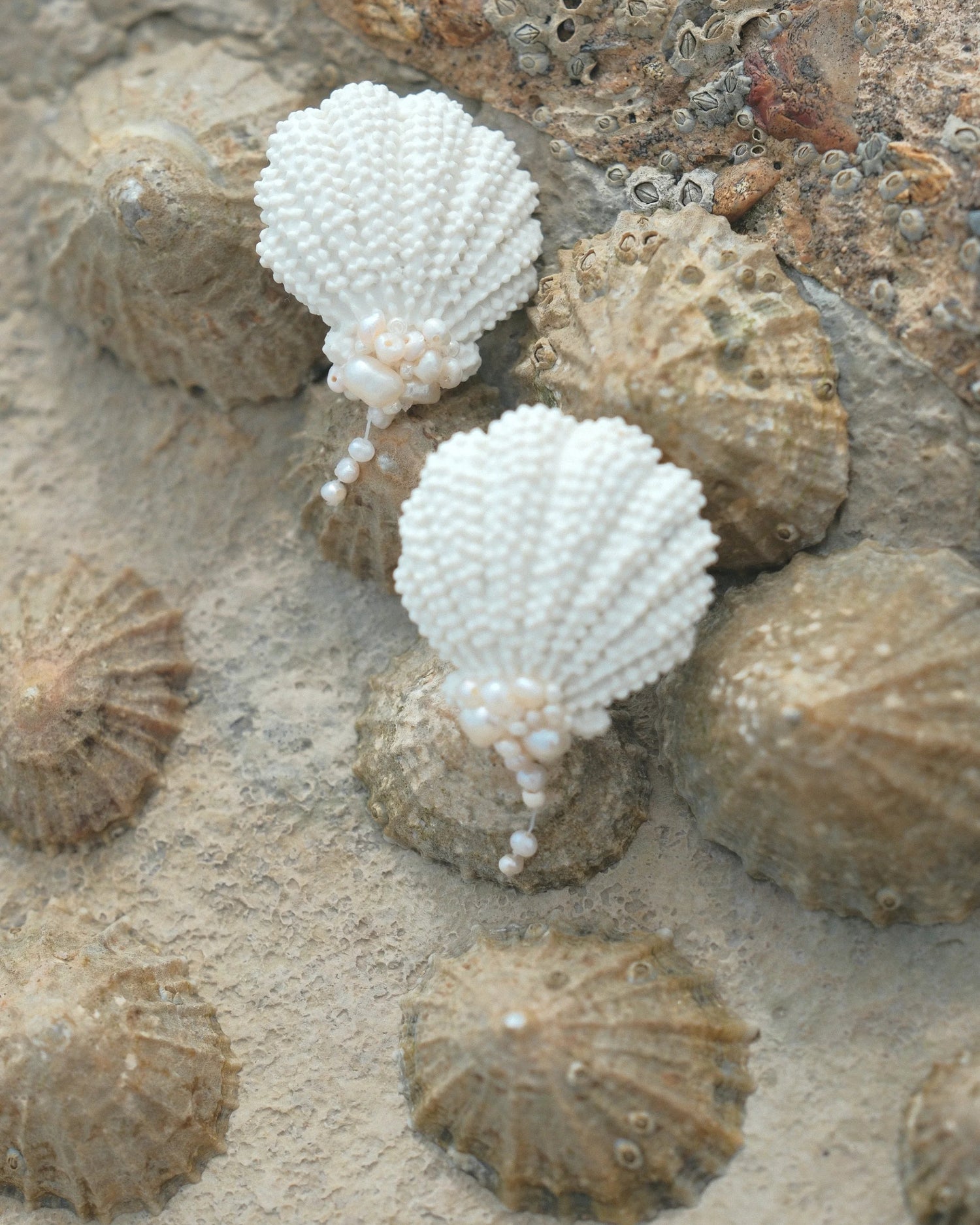 A pair of white beaded shell earrings shaped like scallop shells, resting on a sandy surface surrounded by realistic sea urchin shell props.